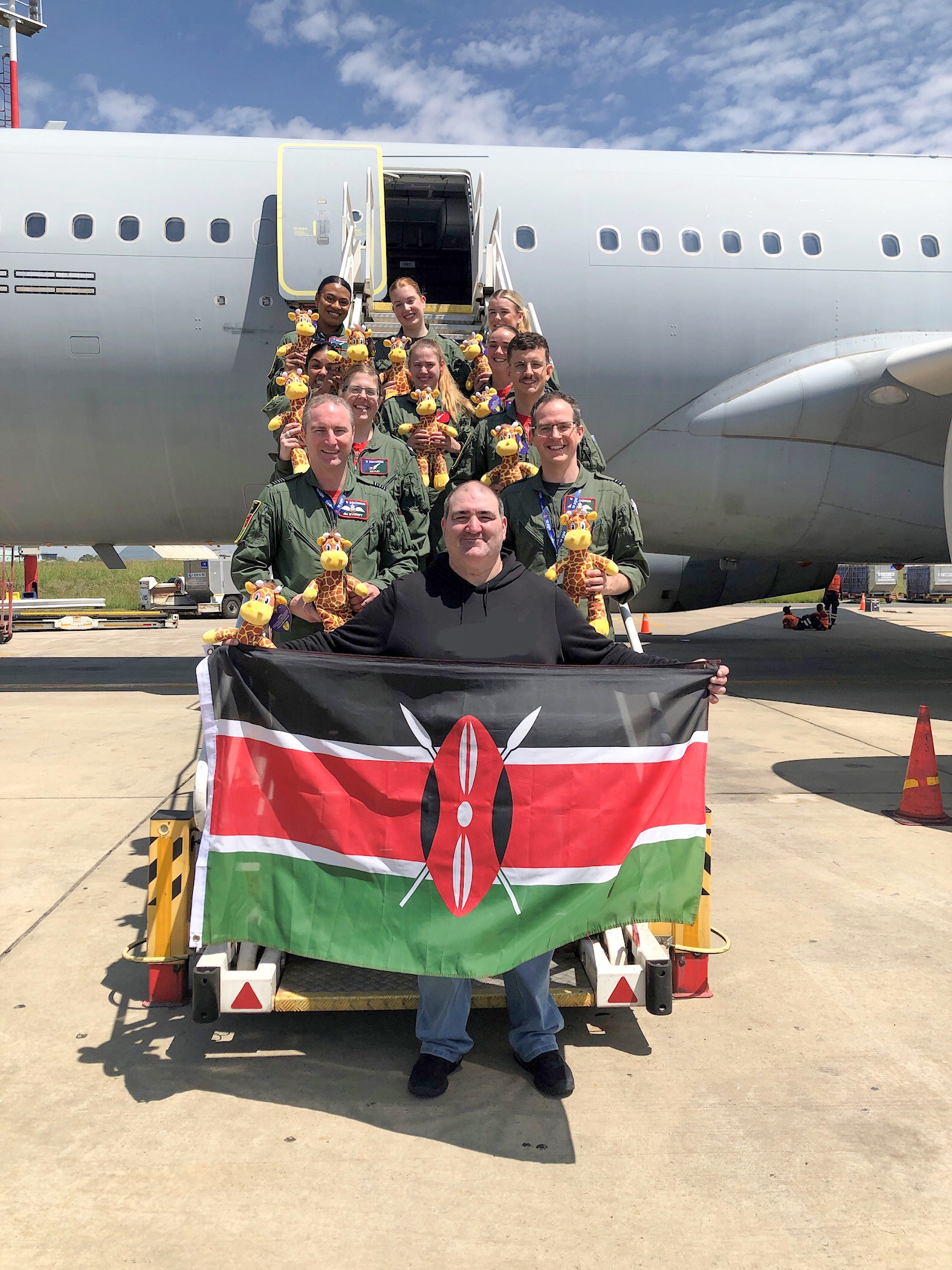 Flight Lieutenant Scott, a pilot on Voyager, and Wing Commander Rycroft, Officer Commanding 10 Squadron, Ian Conway and 8 cabin crew standing on the steps leading to the Voyager passenger door. all are holding a giraffe toy. Ian, who is standing in front is holding an unfurled Kenyan national flag.